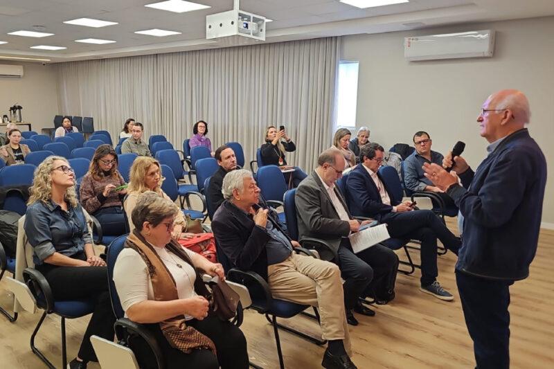 Um palestrante de cabelos grisalhos está em pé, segurando um microfone e falando para uma plateia sentada em cadeiras de auditório. A sala é iluminada e possui paredes claras, com cortinas ao fundo. As pessoas na plateia estão prestando atenção ao palestrante, algumas fazendo anotações ou usando celulares para registrar o momento. A maioria está sentada em cadeiras azuis e veste roupas formais e casuais.