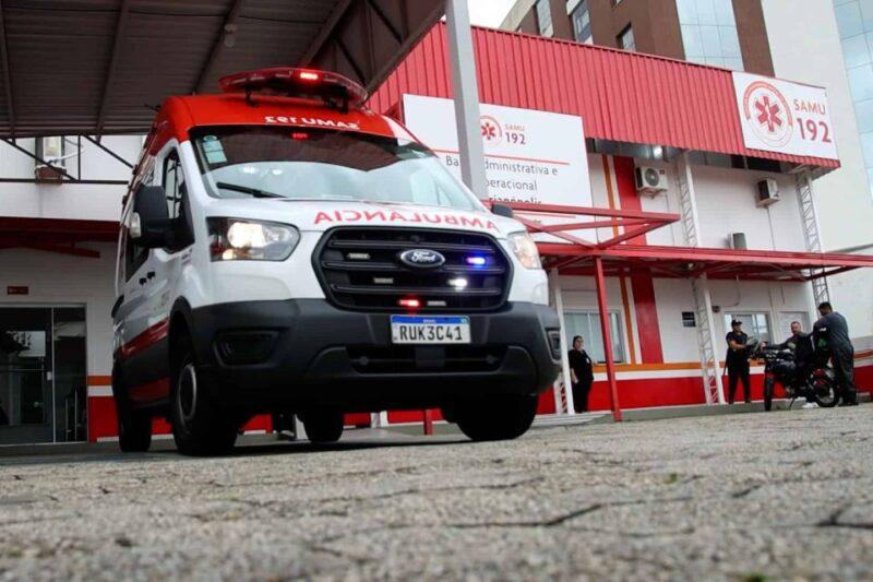 Ambulância do SAMU estacionada em frente a uma base administrativa e operacional. O veículo branco com detalhes vermelhos está de faróis acesos, pronta para uma possível emergência. Ao fundo, parte da equipe de socorristas e um edifício com a logo do SAMU 192 destacada.