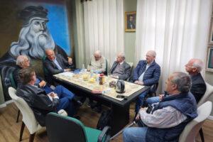 "Imagem de sete homens de meia-idade e idosos sentados ao redor de uma mesa de reunião em uma sala de conferências. Todos estão voltados uns para os outros, participando de uma discussão. A mesa está coberta com uma toalha estampada e contém diversos itens como garrafas de água, xícaras, um bule e alguns recipientes com alimentos. No fundo, à esquerda, há um grande mural na parede retratando um homem com barba longa e chapéu, lembrando uma figura renascentista. Na parede do fundo, à direita, há um retrato emoldurado de outro homem."