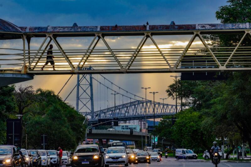 "Imagem de uma passarela para pedestres com estrutura de metal sobre uma avenida movimentada, ao entardecer. Na passarela, uma pessoa está caminhando, criando uma silhueta contra o céu. Abaixo, há vários carros transitando na avenida. Ao fundo, pode-se ver a icônica Ponte Hercílio Luz, um símbolo de Florianópolis, com sua estrutura metálica suspensa. O céu está parcialmente nublado, com raios de sol atravessando as nuvens, criando um efeito dramático de luz e sombra. Árvores verdes flanqueiam a estrada e completam a cena urbana."