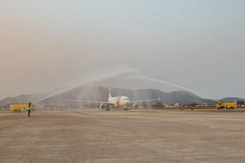 "Avião da TAP Air Portugal no aeroporto durante cerimônia de batismo com jatos d'água, realizado por dois caminhões de bombeiros amarelos. O avião está centralizado na imagem, com os jatos d'água formando um arco sobre ele. Ao fundo, é possível ver montanhas e construções ao longe. Um funcionário do aeroporto, vestindo um colete refletivo, está à esquerda, sinalizando na pista."