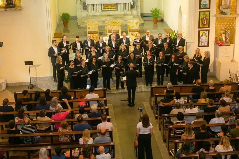 Fotografia de um coral se apresentando dentro de uma igreja. Aproximadamente trinta coralistas, todos vestidos de preto, estão posicionados em três fileiras, segurando partituras. À frente do grupo, um maestro de costas rege a apresentação. O público, sentado em bancos de madeira, observa atentamente. O altar da igreja aparece ao fundo, com uma decoração simples, incluindo plantas e algumas imagens religiosas nas paredes laterais. A iluminação é amarelada e aconchegante, destacando o ambiente solene e reverente.