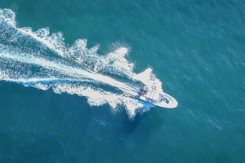 Fotografia aérea de um barco pequeno navegando em alta velocidade em um mar de águas azul-esverdeadas. O barco deixa uma longa esteira de espuma branca na água, que se expande em ondas suaves. O cenário é amplo, com o barco sendo o único elemento visível em meio ao oceano.