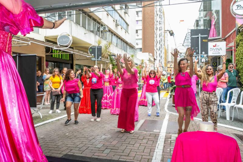 Fotografia de um grupo de mulheres participando de uma atividade ao ar livre em uma rua urbana. Todas estão vestindo roupas predominantemente rosas, com algumas usando tons de vermelho. Elas estão com os braços levantados, como se estivessem seguindo uma coreografia, lideradas por alguém fora da imagem à esquerda. Ao fundo, mais pessoas assistem, algumas vestidas com trajes rosa brilhantes, semelhantes a vestidos de festa. A rua está cercada por prédios e lojas, com algumas cadeiras de plástico brancas à direita.