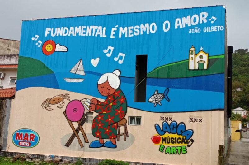 Mural colorido em uma parede de prédio mostra uma cena litorânea. No centro, uma senhora de pele clara e cabelos brancos, vestida com um vestido verde estampado com flores vermelhas, faz renda sentada em um banquinho, com o mar e colinas ao fundo. Ao lado dela, há um caranguejo sorridente e um peixe no mar. No topo, está escrito “Fundamental é mesmo o amor – João Gilberto”, com notas musicais e um sol sorridente ao lado de uma nuvem. No canto inferior direito, o logotipo "Lagoa Música e Arte" e no canto inferior esquerdo o logotipo "Mar Tintas".