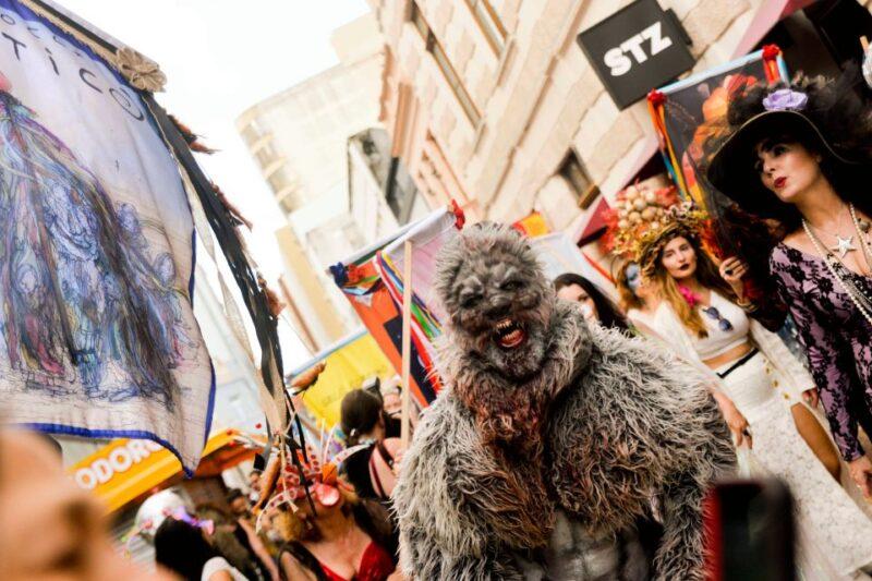 Fotografia de uma festa de rua com pessoas fantasiadas. No centro, uma pessoa vestida como um lobisomem, com máscara realista e traje peludo cinza, caminha em direção à câmera. À direita, uma mulher com um vestido rendado preto e um grande chapéu decorado também participa da celebração. Ao fundo, outras pessoas estão fantasiadas, e há bandeiras e enfeites coloridos segurados por participantes, criando um ambiente vibrante e festivo. A festa ocorre em uma rua cercada por prédios antigos.