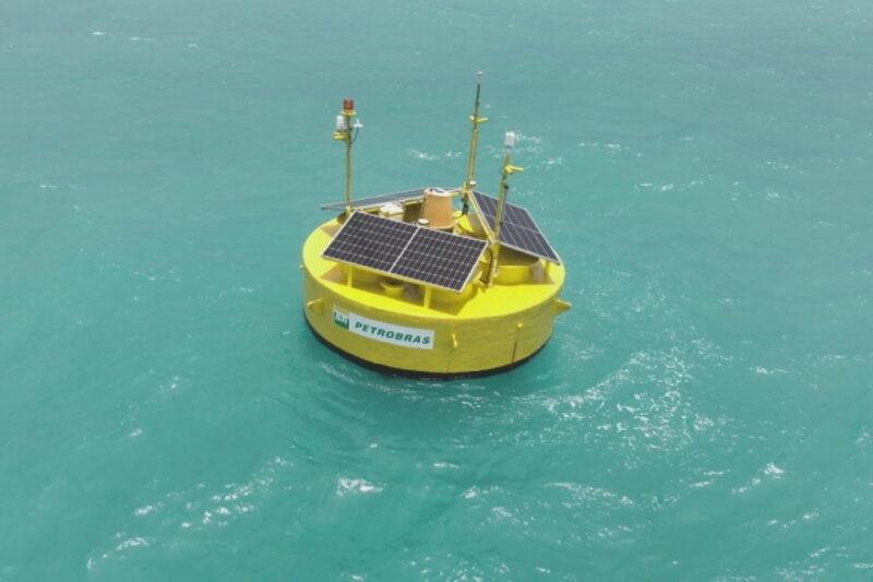 Bóia flutuante amarela com placas solares em sua superfície, equipada com sensores e antenas, flutua em um mar de cor azul-turquesa. A bóia exibe o logotipo da Petrobras, indicando ser parte de um projeto de monitoramento ambiental ou oceanográfico da empresa.