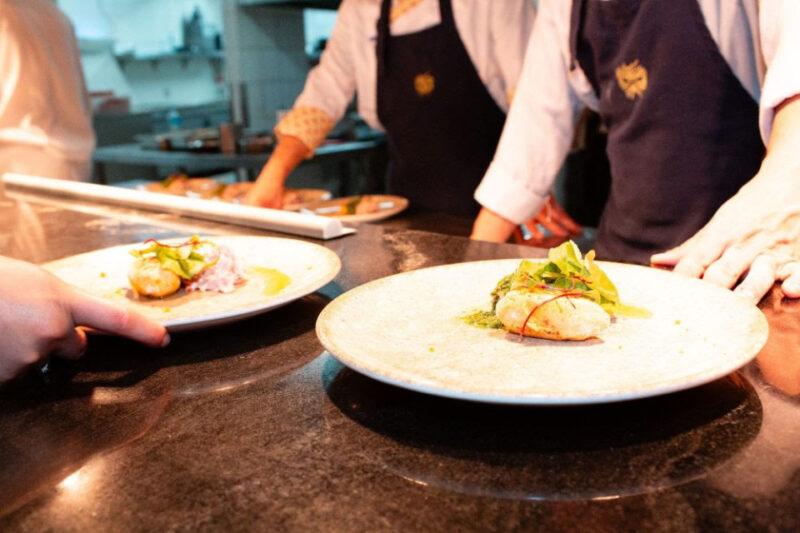 Pratos gourmet recém-preparados são apresentados em uma cozinha profissional. Em primeiro plano, dois pratos de cerâmica contêm uma porção de comida delicadamente montada com folhas verdes como guarnição. Ao fundo, chefs de cozinha com uniformes brancos e aventais escuros estão preparando ou supervisionando os pratos, evidenciando um ambiente de culinária sofisticada e artesanal