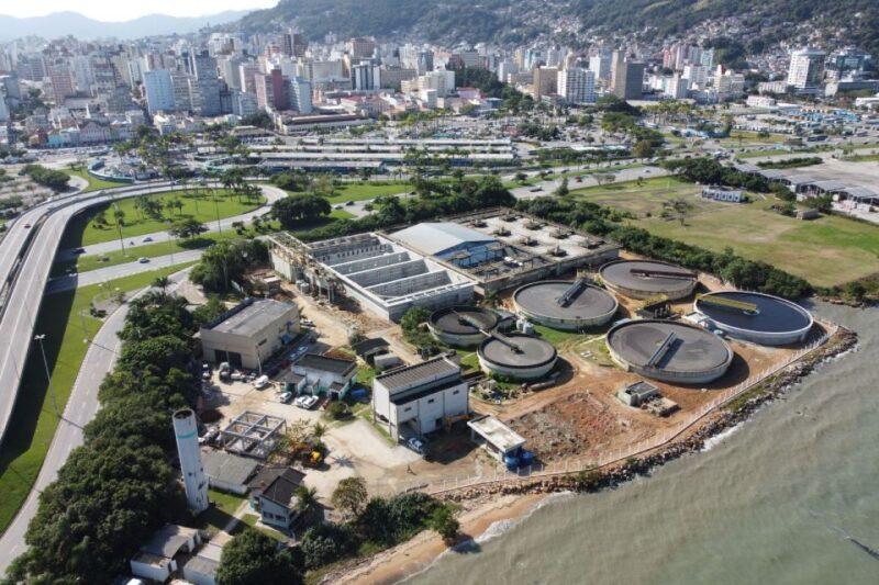 Foto aérea de uma estação de tratamento de esgoto localizada à beira-mar. A instalação possui vários tanques circulares grandes, dispostos ao lado de edifícios industriais retangulares. Ao fundo, há uma rodovia de múltiplas faixas à esquerda, além de áreas verdes e uma grande área urbana com muitos prédios altos. O mar aparece na parte inferior direita da imagem, e colinas cobertas de vegetação estão ao fundo, atrás da cidade.