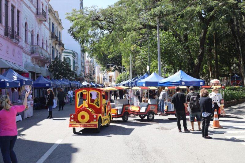 Em uma rua aberta para pedestres, várias pessoas caminham ao redor de barracas azuis de feira, montadas ao longo da calçada à esquerda, em frente a prédios antigos de arquitetura colorida, como fachadas rosa e verde. No centro da imagem, há um trenzinho turístico infantil, vermelho e amarelo, com a inscrição "Viagem no Tempo" na parte traseira. Algumas pessoas estão próximas ao trenzinho, enquanto uma mulher à esquerda, de blusa rosa, acena. Árvores grandes cercam o lado direito da rua, criando uma atmosfera de feira ao ar livre em um dia ensolarado.