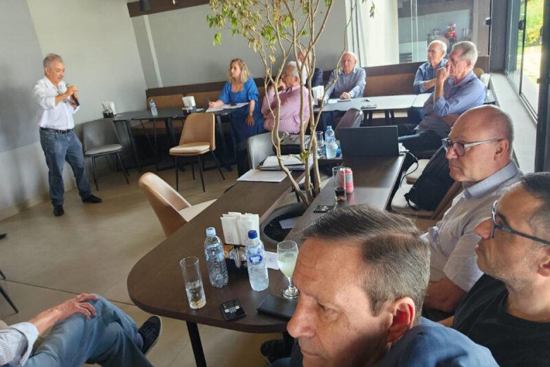 Fotografia de uma reunião em uma sala com mesas de madeira e cadeiras em tom claro. À esquerda, um homem em pé segura um microfone enquanto fala para os participantes sentados ao redor das mesas. No centro, há um pequeno vaso com uma planta. A maioria das pessoas aparenta estar prestando atenção, e há garrafas de água, copos e objetos pessoais sobre as mesas. A sala tem iluminação natural, vinda de janelas amplas à direita. O ambiente sugere um encontro formal ou profissional.