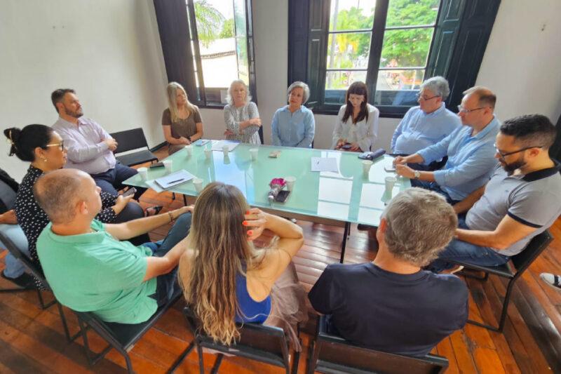 Em uma sala iluminada com piso de madeira e uma janela grande ao fundo, onze pessoas estão reunidas ao redor de uma mesa retangular de vidro. Elas estão sentadas em cadeiras, formando um círculo, e parecem atentas e engajadas em uma discussão. Sobre a mesa, há folhas de papel, copos descartáveis e uma pequena flor em embalagem, indicando um ambiente de reunião formal. Ao fundo, pela janela, é possível ver árvores e parte de uma área externa iluminada.