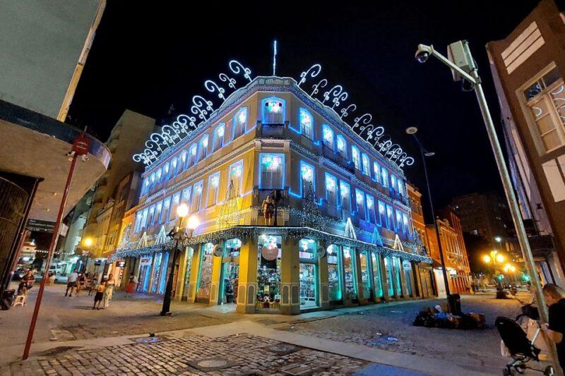 Fotografia noturna de uma rua com um prédio histórico de três andares decorado com luzes natalinas. O edifício, de cor amarela, está iluminado por contornos de luzes azuis ao redor das janelas, e ornamentos brancos no telhado, que se destacam contra o céu escuro. Pequenas árvores decorativas e guirlandas de luzes complementam a fachada. À esquerda, algumas pessoas caminham pela rua de paralelepípedos, enquanto à direita, um homem empurra um carrinho de bebê. As luminárias de rua também estão acesas, criando um ambiente festivo e acolhedor.