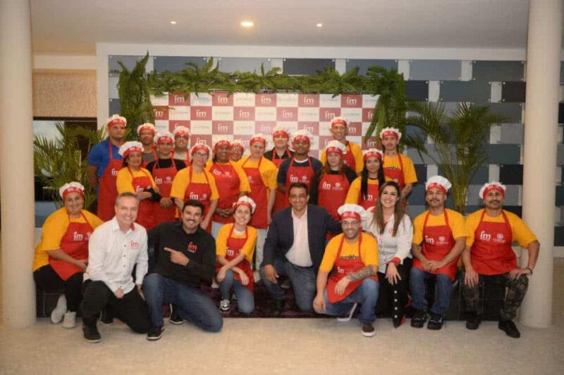 Fotografia de um grupo de vinte pessoas posando para uma foto em um ambiente interno. A maioria usa aventais vermelhos e camisetas amarelas, além de toucas de cozinheiro brancas e vermelhas, sugerindo que participam de um evento culinário ou workshop. Na frente do grupo, quatro pessoas estão agachadas ou ajoelhadas, duas delas vestindo roupas casuais (uma delas com camiseta preta apontando para o lado, e a outra com camisa social branca). Ao fundo, há um painel com o logotipo "IM" repetido e plantas decorativas. O ambiente parece ser de um evento de participantes de gastronomia.