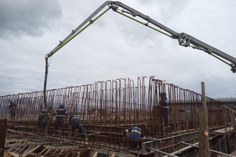 Obra em construção com operários trabalhando em uma estrutura de ferro armado. Há diversas barras de aço verticais e horizontais formando uma armação, e no alto da imagem, um braço mecânico de bomba de concreto aponta para a estrutura. O céu está nublado, criando uma atmosfera cinzenta. Os trabalhadores vestem uniformes de segurança com faixas refletivas.