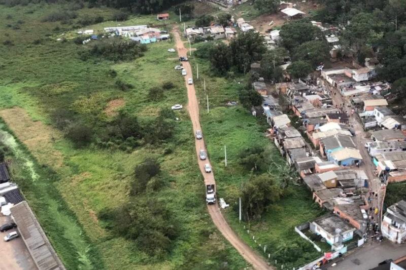 Imagem aérea de uma estrada de terra que atravessa uma área verde com pouca vegetação, conectando uma vila de casas simples à direita da imagem. Há vários carros alinhados ao longo da estrada, alguns se aproximando da vila. À esquerda, a área verde é ampla e desocupada, enquanto à direita a vila mostra casas construídas próximas umas das outras, com pessoas espalhadas pelas ruas. Ao fundo, vê-se mais vegetação e construções dispersas.