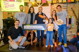 Fotografia de um grupo de seis pessoas sorridentes em um evento de premiação. No centro, um homem segura um troféu e uma garrafa de espumante, com uma faixa escrita "primeiro CAMPEÃO FLORIANÓPOLIS". Ao lado dele, uma mulher segura uma criança, que exibe um certificado laranja de campeã. Duas pessoas à esquerda vestem camisetas cinzas com a frase "BUTECO COM 'U'", e uma mulher à direita segura um prato decorativo com o logo "Comida di Buteco". Ao fundo, há um painel com o texto "SOMOS TODOS BUTECO - edição dois mil e vinte e quatro", em um ambiente com paredes de tijolos aparentes.