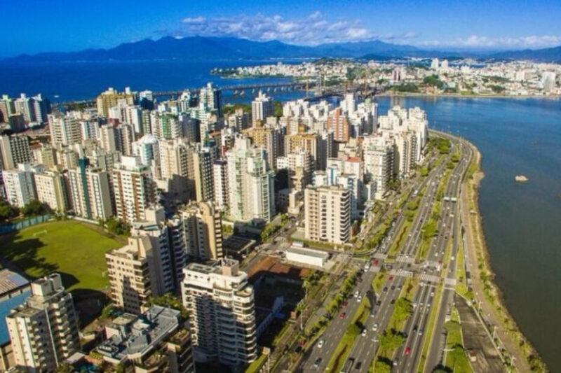 Imagem aérea de uma área urbana à beira-mar, com muitos prédios altos e modernos ao longo de uma avenida movimentada. À direita, uma ampla faixa de água separa a cidade de outra área ao fundo, com montanhas e céu azul. O cenário mostra uma combinação de urbanização densa com áreas naturais ao redor.