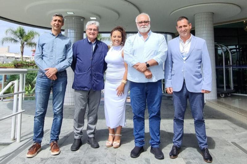 Cinco pessoas posam lado a lado em frente a um prédio com colunas revestidas de azulejos brancos. Da esquerda para a direita: um homem de camisa azul claro e jeans, um homem de jaqueta azul escura, uma mulher de vestido branco, um homem de camisa azul clara e jeans, e um homem de blazer azul claro sobre camisa branca. Eles sorriem, em um ambiente externo e iluminado, com plantas e edifícios ao fundo.