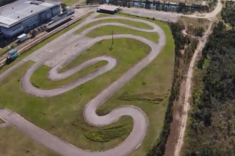 Imagem aérea de uma pista sinuosa de kart, localizada em uma área gramada e cercada por vegetação. A pista tem várias curvas fechadas e traçado em forma de "S". À esquerda, há um grande edifício industrial, e ao fundo, um pequeno lago e áreas de mata. Um caminho de terra corre ao lado direito da imagem, conectando a pista a outras áreas do local.