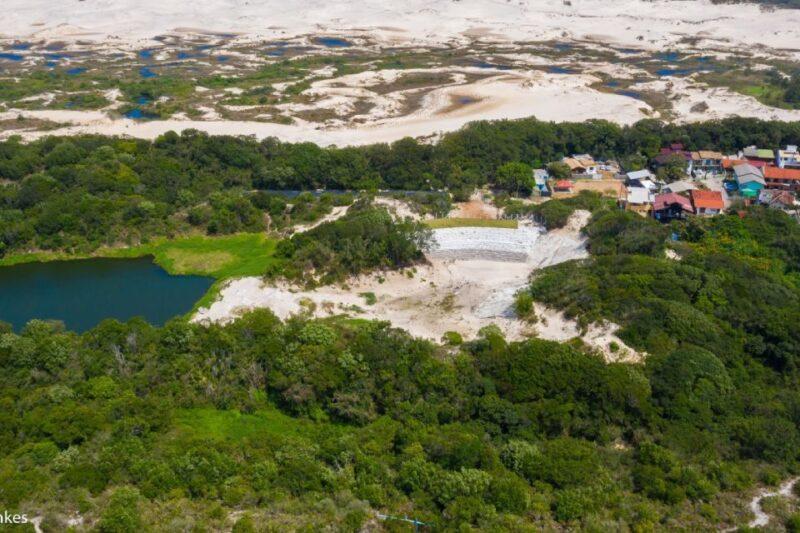 Imagem aérea de uma área de preservação com vegetação verde densa, um lago de águas escuras e, ao fundo, uma extensão de dunas de areia clara. No canto superior direito, vê-se um pequeno aglomerado de casas coloridas ao lado da área verde, sugerindo uma comunidade próxima às dunas. A vegetação é interrompida por uma clareira com uma estrutura branca em formato de escadaria, que parece uma intervenção artificial na paisagem natural.