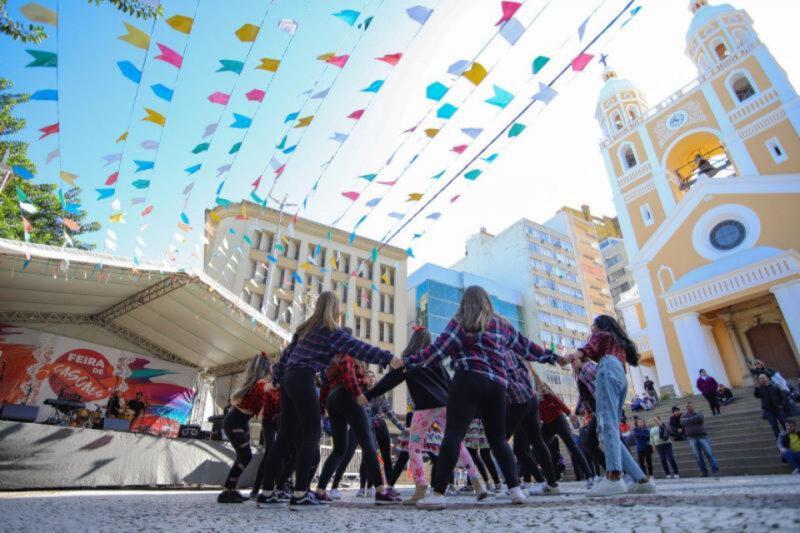 Fotografia de uma apresentação ao ar livre durante um evento festivo. Um grupo de jovens, a maioria vestindo camisas xadrez e calças escuras, dança em círculo no centro da imagem. Acima deles, bandeirinhas coloridas estão penduradas, estendendo-se por todo o espaço. À esquerda, há um palco com músicos e um grande painel de fundo com o texto "Feira de Cascaes". À direita, uma igreja amarela com duas torres e um relógio é vista ao fundo, com algumas pessoas sentadas nas escadas em frente. O céu está claro, com prédios ao redor compondo a paisagem urbana.