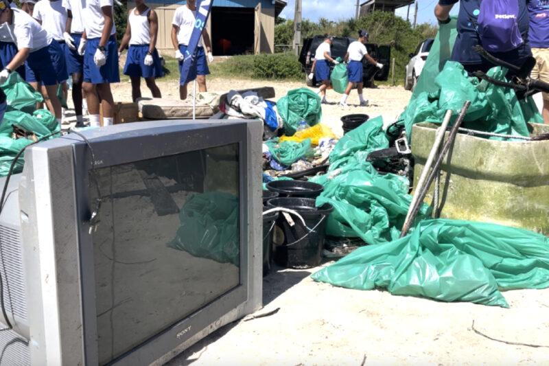 Grupo de jovens vestindo uniformes brancos e azuis participa de uma ação de limpeza em uma área de praia. No chão, há uma televisão de tubo e diversos sacos verdes grandes cheios de lixo, além de baldes e pedaços de madeira e metal. Ao fundo, algumas pessoas carregam sacos enquanto outras observam a área, onde também há veículos e uma pequena construção em madeira.