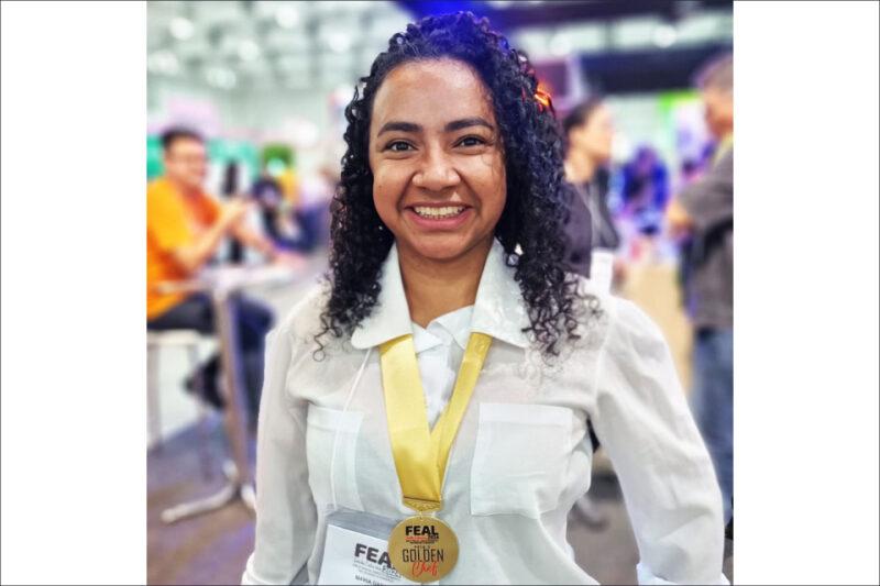 Fotografia de uma mulher sorridente de cabelos cacheados e pretos, usando uma camisa branca e uma medalha dourada pendurada no pescoço. A medalha tem os dizeres “FEAL Golden Chef dois mil e vinte e três”. Ela está em um ambiente interno com pessoas desfocadas ao fundo, que parecem estar participando de um evento.