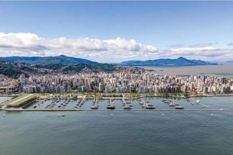 Vista aérea panorâmica da orla da cidade de Florianópolis, destacando uma marina com barcos atracados em diferentes píeres. Em primeiro plano, há o mar em tons de azul e verde, enquanto a linha costeira é marcada por edifícios altos e modernos ao longo da Avenida Beira-Mar Norte. Ao fundo, montanhas cobertas de vegetação se estendem até o horizonte, sob um céu azul com algumas nuvens brancas dispersas. A paisagem reflete uma combinação de urbanização e natureza, mostrando a harmonia entre a área costeira e a cidade.