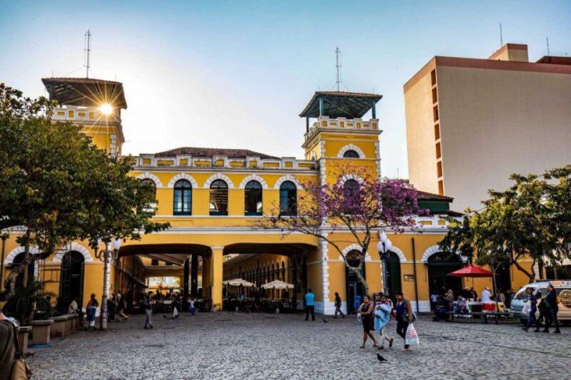 Fotografia de um mercado público com fachada amarela e detalhes brancos, composta por arcos e janelas no andar superior. No centro, há uma passagem coberta, onde se veem pessoas caminhando e mesas com guarda-sóis. A praça em frente é de paralelepípedos e conta com árvores, sendo uma delas com flores roxas. À direita, algumas pessoas estão sentadas em mesas e há um quiosque vermelho. Ao fundo, um edifício alto bege contrasta com o céu claro e azul.