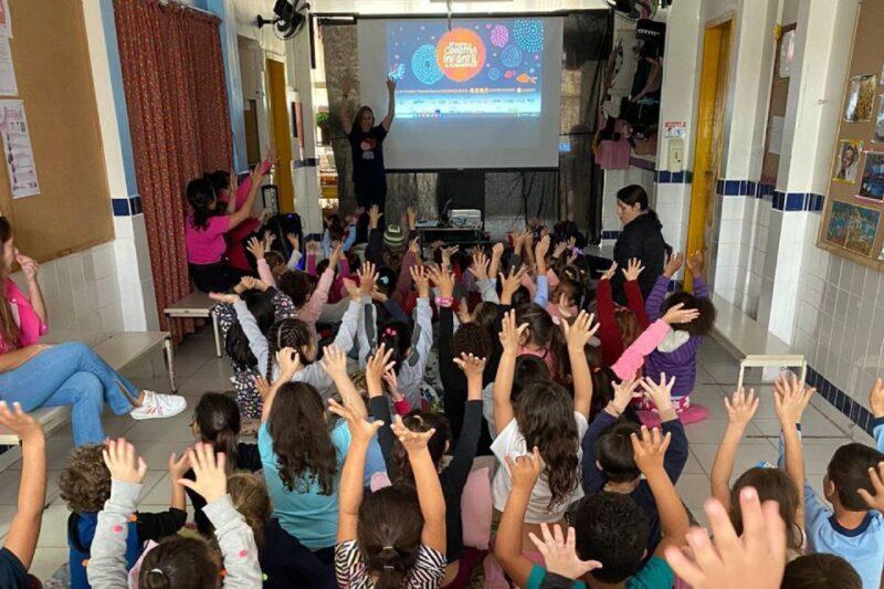 Em uma sala com paredes brancas e detalhes em azul, várias crianças estão sentadas no chão, todas com as mãos levantadas. Elas estão voltadas para uma tela de projeção, onde aparece uma apresentação com o título "Cinema Infantil". Há duas mulheres presentes, uma à esquerda, sentada, e outra de pé ao fundo, com os braços levantados, provavelmente conduzindo a atividade. A sala é decorada com painéis nas paredes e alguns materiais de papel. A atmosfera é de entusiasmo, com as crianças participando ativamente.