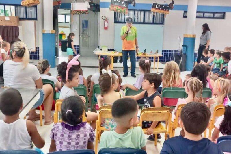 Fotografia de uma atividade educativa em uma sala de aula com várias crianças sentadas em cadeiras coloridas, voltadas para um homem em pé, que fala ao microfone. Ele usa uma camisa verde e laranja e um boné. À sua frente, há uma mesa com objetos diversos. Algumas crianças estão atentas, enquanto outras conversam ou olham ao redor. No fundo, uma parede azul e branca, com decorações suspensas e uma mesa ao lado, complementa o ambiente. Uma mulher de pé, à direita, observa a atividade.