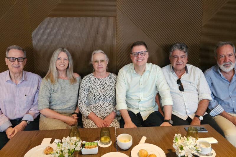 Seis pessoas estão sentadas lado a lado em uma mesa de madeira. Da esquerda para a direita: um homem de óculos, com cabelo grisalho, usando camisa social listrada; uma mulher loira de cabelo solto, com blusa cinza clara; uma mulher de óculos com cabelo grisalho preso, vestindo um vestido branco com estampa preta; um homem sorridente de óculos, com cabelo curto, vestindo uma camisa branca de mangas dobradas; um homem de cabelo grisalho, usando camisa clara e óculos escuros pendurados na gola; e outro homem de barba branca, vestindo uma camisa azul clara. Sobre a mesa, há xícaras, pratos com pequenos pães, flores brancas e copos com bebidas.