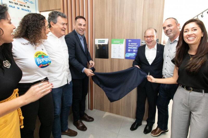 Fotografia de um grupo de seis pessoas sorrindo durante uma inauguração. No centro, dois homens seguram um pano azul, prestes a revelar uma placa em uma parede de madeira. A pessoa à esquerda usa uma camiseta branca com o logo "Festival Viva Floripa", e outras estão vestidas com roupas casuais ou formais. A parede atrás deles tem alguns cartazes, incluindo um que diz "A nossa força é o seu negócio" e outro com o logo do Sebrae. Todos parecem estar celebrando o momento.