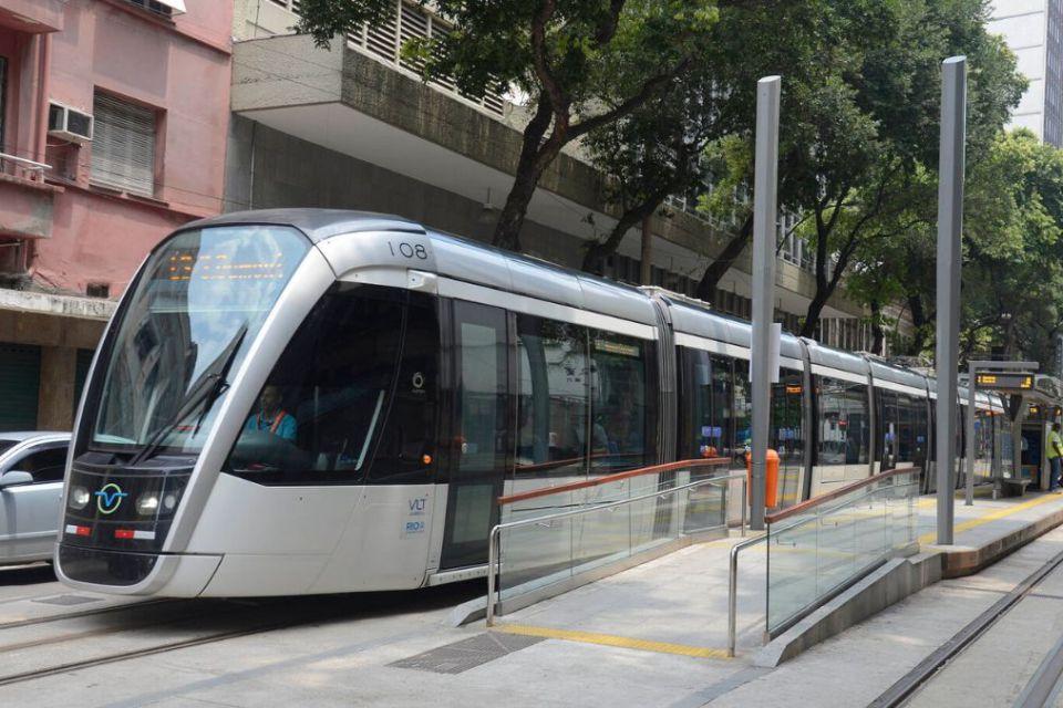 Fotografia de um VLT (Veículo Leve sobre Trilhos) parado em uma estação urbana. O VLT tem design moderno, com frente arredondada e grandes janelas de vidro. Sua cor predominante é prata, com detalhes em preto e azul. A estação está vazia, com algumas pessoas sentadas ao fundo à espera do transporte. Árvores e edifícios residenciais cercam a rua, e um carro prata está estacionado à esquerda. O ambiente é urbano, com clima de rotina diária.