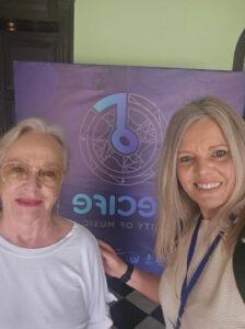 Fotografia de duas mulheres sorrindo, posando lado a lado em frente a um banner roxo com o texto parcialmente visível "RECIFE - City of Music" e um logotipo circular estilizado com uma nota musical. A mulher à esquerda é mais velha, tem cabelos grisalhos presos e usa óculos de sol e camiseta branca. A mulher à direita tem cabelos loiros soltos, veste uma blusa clara e um crachá azul no pescoço.