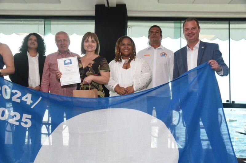 Grupo de seis pessoas posando lado a lado em ambiente interno, com uma grande bandeira azul estendida na frente, onde se lê "dois mil e vinte e quatro / dois mil e vinte e cinco" e se vê o símbolo da *bandeira azul*, uma certificação internacional de qualidade ambiental. Ao centro, um homem idoso segura um certificado, ao lado de uma mulher de vestido estampado. A mulher ao centro está vestida de branco e sorri para a foto, assim como o homem de terno à direita que segura a bandeira.