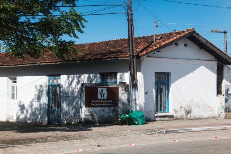 Fotografia de uma construção histórica com paredes brancas e telhado de telhas coloniais avermelhadas, identificada como "Casarão Aéropostale". Na fachada, há uma placa marrom com o texto "Casarão Aéropostale - Patrimônio Histórico" e um logotipo de um avião antigo. As janelas e portas possuem grades azuis, e na calçada, ao lado de um poste, estão sacos verdes de lixo.