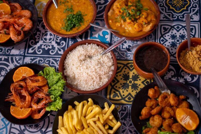 Fotografia de uma mesa repleta de pratos típicos da culinária brasileira, dispostos sobre uma toalha com estampas coloridas. No centro, uma tigela com arroz branco. Ao redor, há um prato com camarões grelhados acompanhados de folhas de alface e fatias de laranja, batatas fritas, camarões empanados com laranja decorativa, uma tigela com molho de feijão, e tigelas com vatapá e moqueca de camarão, ambos decorados com cheiro-verde. Há também um recipiente com farinha de mandioca. A disposição dos pratos cria uma composição vibrante e apetitosa.