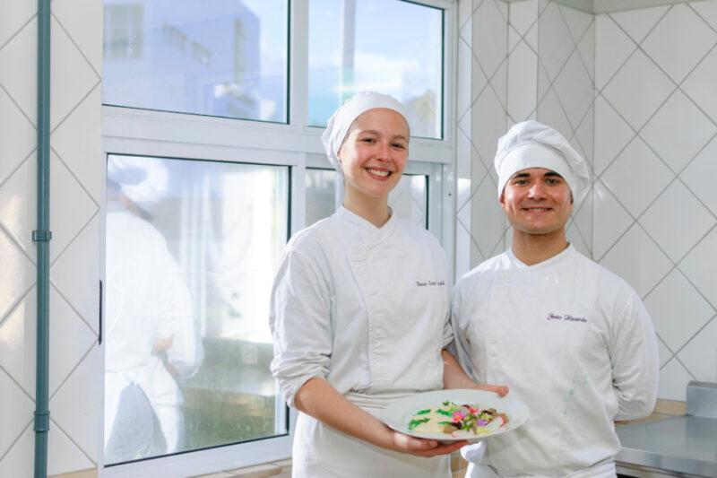 Dois jovens chefs sorrindo em uma cozinha clara, revestida com azulejos brancos. Eles vestem uniformes de chef brancos com toucas, e a mulher à esquerda segura um prato decorado com uma preparação colorida. Ao fundo, uma janela permite a entrada de luz natural. Ambos demonstram satisfação e orgulho.