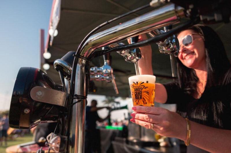 Mulher de óculos escuros e cabelos escuros serve chope em um copo plástico transparente decorado com a inscrição "Ands Craft Beer" e desenhos estilizados. Ela sorri enquanto segura o copo sob a torneira de um equipamento metálico de chopeira. Ao fundo, o ambiente é aberto e iluminado pelo sol, sugerindo um evento ao ar livre.