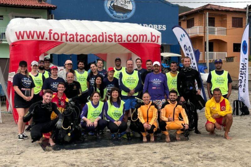 Grupo de cerca de vinte pessoas posando para foto em uma praia, reunidos para uma ação de limpeza. A maioria usa coletes verdes com o logotipo "Limpeza dos Mares" e alguns estão vestidos com roupas de mergulho pretas ou amarelas. Eles estão de pé ou agachados sob um portal inflável vermelho e branco com o endereço "www.fortatacadista.com.br" e ao lado de bandeiras verticais também com o logotipo "Limpeza dos Mares". Ao fundo, há construções simples, incluindo um prédio azul com o letreiro "Pescadores Pântano do Sul".