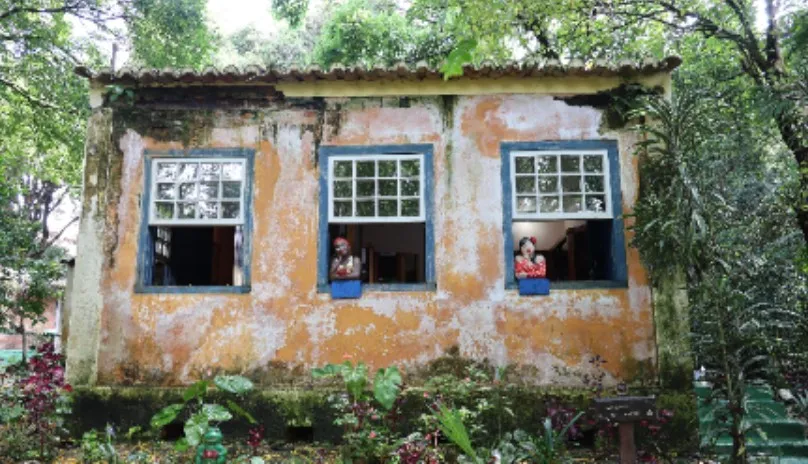 Casa antiga de fachada desgastada com pintura amarela e detalhes azuis ao redor das janelas, localizada em meio a um jardim com plantas variadas. Três janelas estão abertas, e em duas delas há bonecas de cerâmica vestidas com trajes típicos, expostas sobre as janelas como se estivessem olhando para fora. A casa está cercada por árvores, criando um ambiente rústico e acolhedor em meio à natureza.