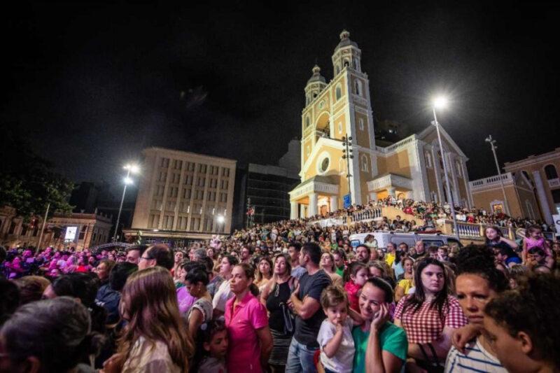 Fotografia noturna de uma grande multidão em frente a uma igreja de arquitetura colonial, com fachadas em tons de amarelo e branco e duas torres. As pessoas, de várias idades, parecem concentradas e aguardam algum evento, olhando para frente. Muitas seguram celulares, e algumas estão acompanhadas de crianças. O ambiente é iluminado por postes e luzes artificiais, destacando o prédio da igreja e a multidão ao redor. Ao fundo, vê-se prédios mais modernos.