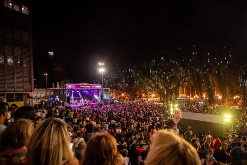 Uma grande multidão assiste a um show noturno ao ar livre em um palco iluminado, com uma faixa escrita “Natal Mágico” no topo. A praça está cheia de pessoas, e árvores decoradas com luzes de Natal criam um ambiente festivo.