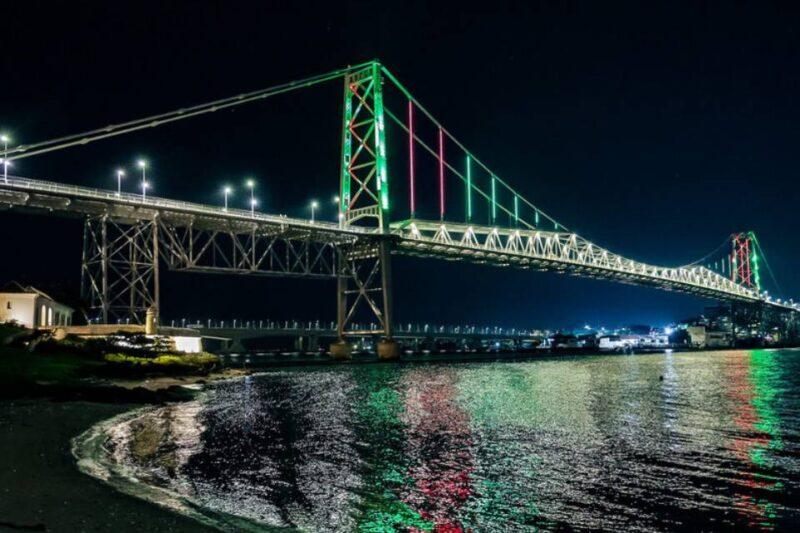 Fotografia noturna de uma ponte metálica iluminada com luzes coloridas nas cores verde, vermelho e branco, refletindo na água abaixo. A estrutura é imponente, com arcos e cabos sustentados por pilares altos. À esquerda, há uma pequena construção iluminada próxima a uma área gramada, enquanto, à direita, são visíveis algumas edificações urbanas ao fundo. O céu está escuro e sem estrelas, destacando a iluminação da ponte e seus reflexos.