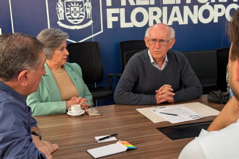 Em uma sala de reuniões da Prefeitura de Florianópolis, quatro pessoas estão sentadas ao redor de uma mesa discutindo um documento. À direita, um homem idoso de cabelos brancos e óculos fala enquanto mantém as mãos unidas sobre a mesa. À esquerda, uma mulher de cabelos curtos grisalhos e blazer verde-claro escuta atentamente, com uma xícara de café e um celular à sua frente. No centro da mesa há papéis, um caderno, uma caneta e marcadores coloridos. Ao fundo, um painel azul com o brasão e o nome "Prefeitura de Florianópolis" em destaque.