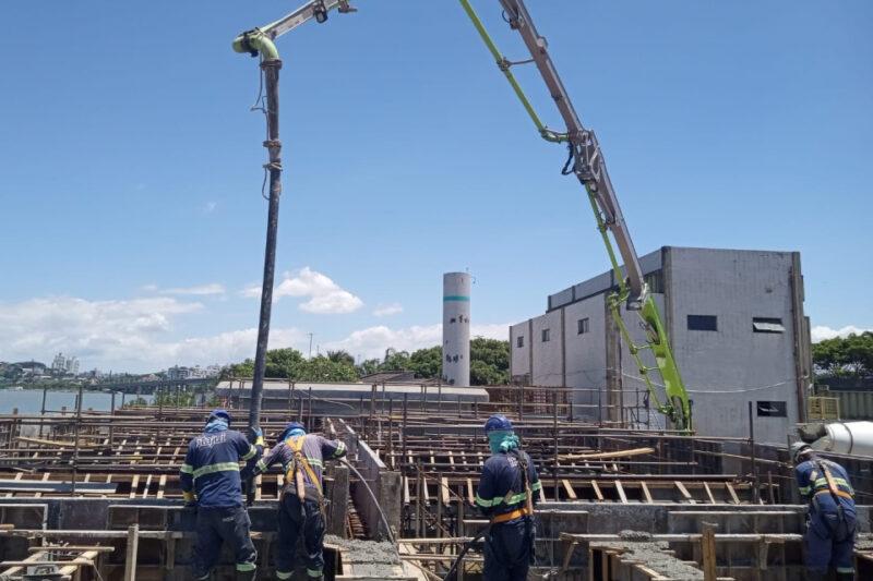 Fotografia de uma construção em andamento em um dia ensolarado. Quatro operários, usando uniformes azuis, capacetes e equipamentos de segurança, trabalham sobre uma estrutura de madeira e aço. No centro, uma máquina com braço articulado verde despeja concreto através de um tubo preto. Ao fundo, é possível ver um edifício cinza, uma torre cilíndrica branca com detalhes azuis, árvores e parte de um rio, além de construções urbanas ao longe. O céu está limpo, com poucas nuvens.