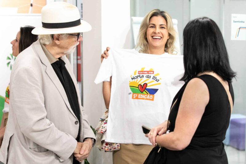 Em uma fotografia colorida, uma mulher loira ao centro sorri enquanto segura uma camiseta branca com a estampa colorida que diz "Festival Viva Norte da Ilha - Quinta Edição" junto a um coração e elementos gráficos de sol. Ela está cercada por duas pessoas: à esquerda, um homem de chapéu branco, óculos escuros e terno claro, que observa a camiseta, e à direita, uma mulher de cabelos escuros usando um vestido preto. Ao fundo, outras pessoas e partes do ambiente de um local interno são visíveis, sugerindo um evento ou encontro.