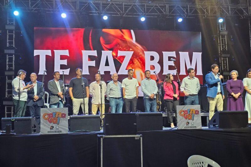 Fotografia de um palco durante um evento oficial, com um grupo de aproximadamente 13 pessoas alinhadas lado a lado. Elas estão em pé, algumas segurando microfones, enquanto um homem à direita está discursando. O fundo do palco exibe um painel luminoso com os dizeres "TE FAZ BEM" em letras brancas. Na parte inferior, cartazes indicam o "Carnaval 2025" como temática do evento. A iluminação do palco é forte, com refletores e estruturas metálicas visíveis. As roupas variam entre trajes formais e casuais, e o clima aparenta ser de celebração.